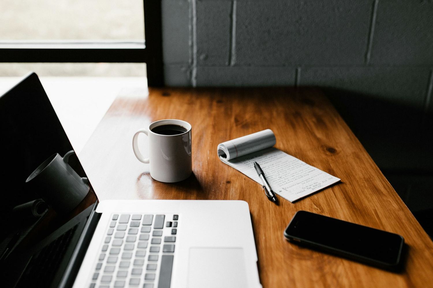 An open laptop, a notebook, and a coffee mug on a wooden desk.