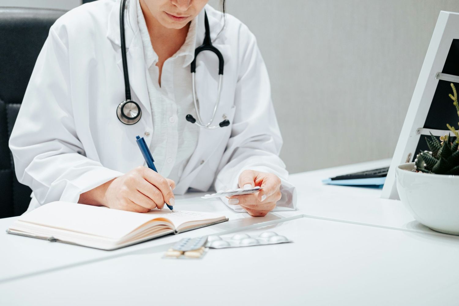 A clinician in a white coat with a stethoscope writing notes at a desk.