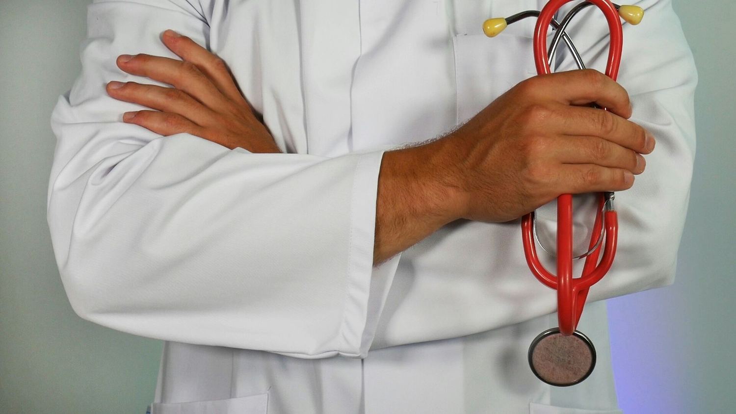 A person in a white coat holding a red stethoscope, arms crossed.