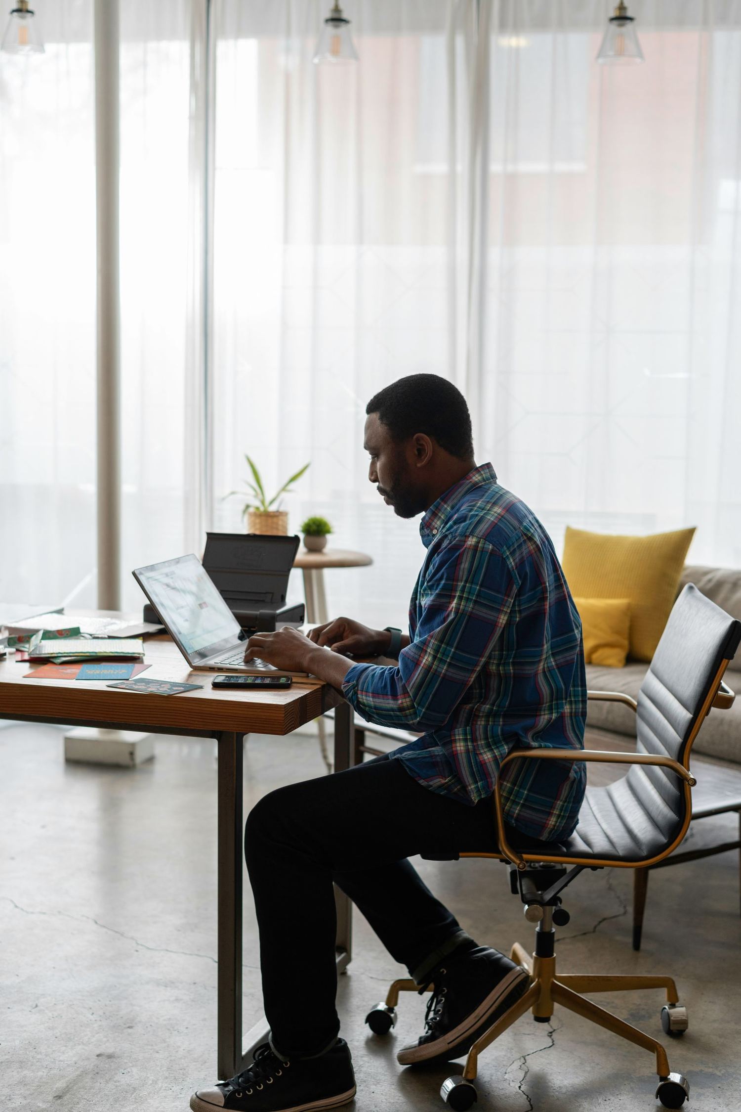 A man working on a laptop at a desk by tall windows.