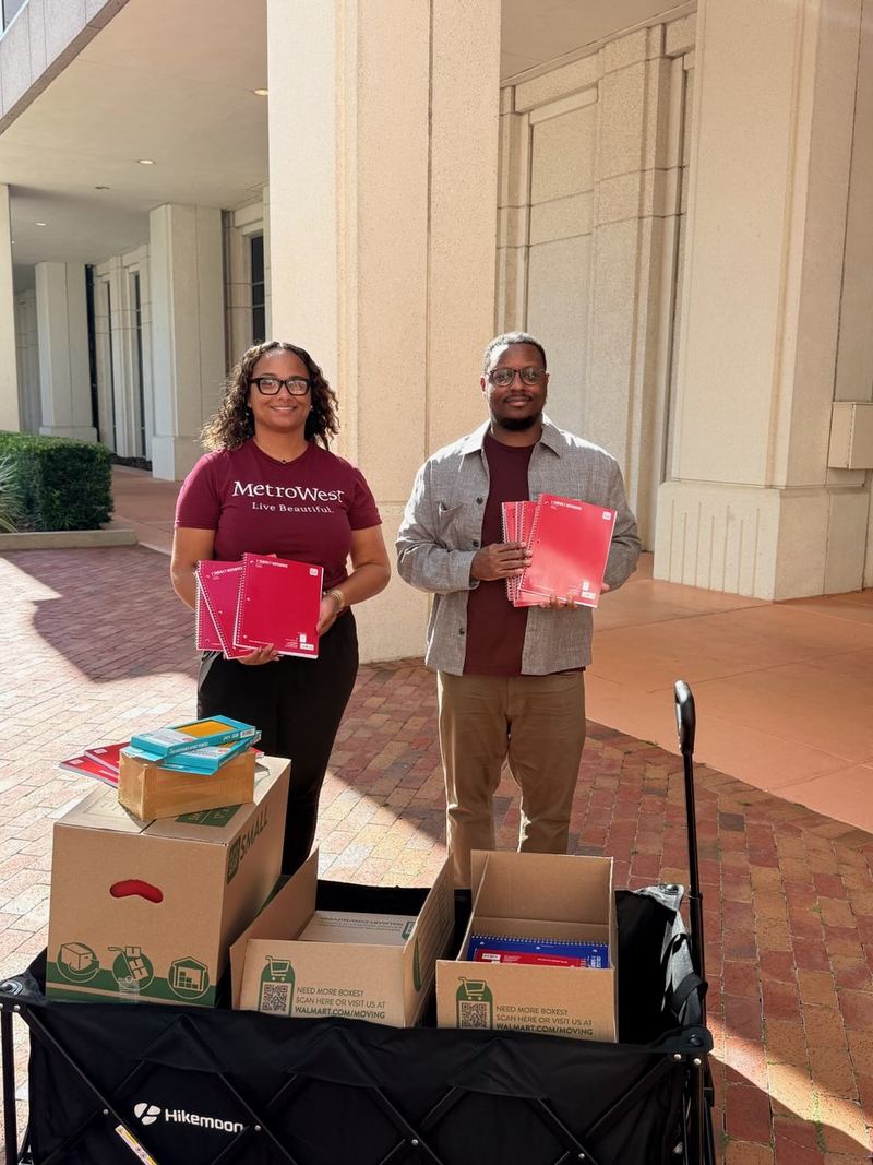 Derrick Johnson and a MetroWest community partner holding notebooks beside a wagon of school supplies