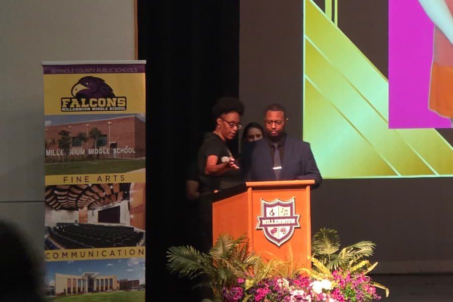 Derrick Johnson speaking at a lectern on stage at a Seminole County Public Schools event at Millennium Middle School, students in the foreground