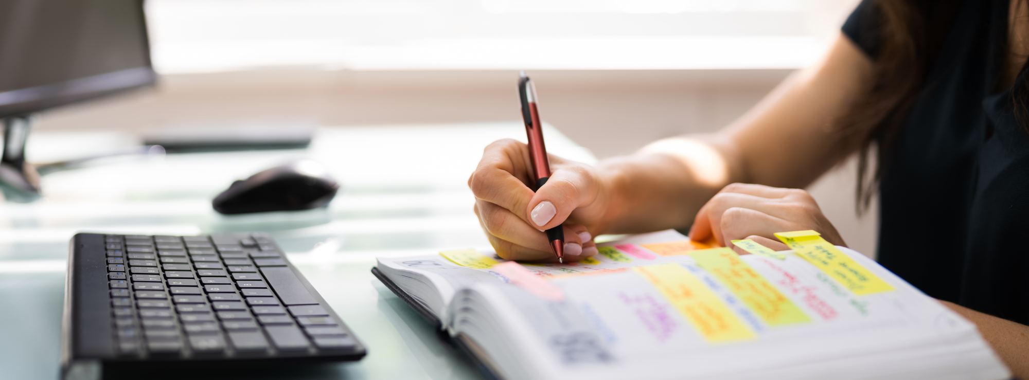 A hand writing notes in a planner beside a keyboard